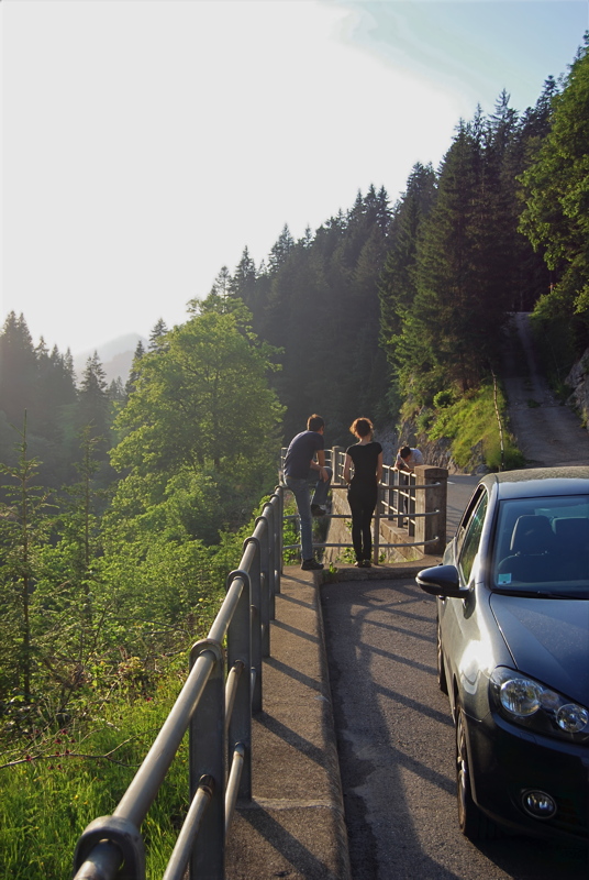 20 - A scenic mountain road overlooking a forested valley with a parked car and people leaning on a concrete retaining wall